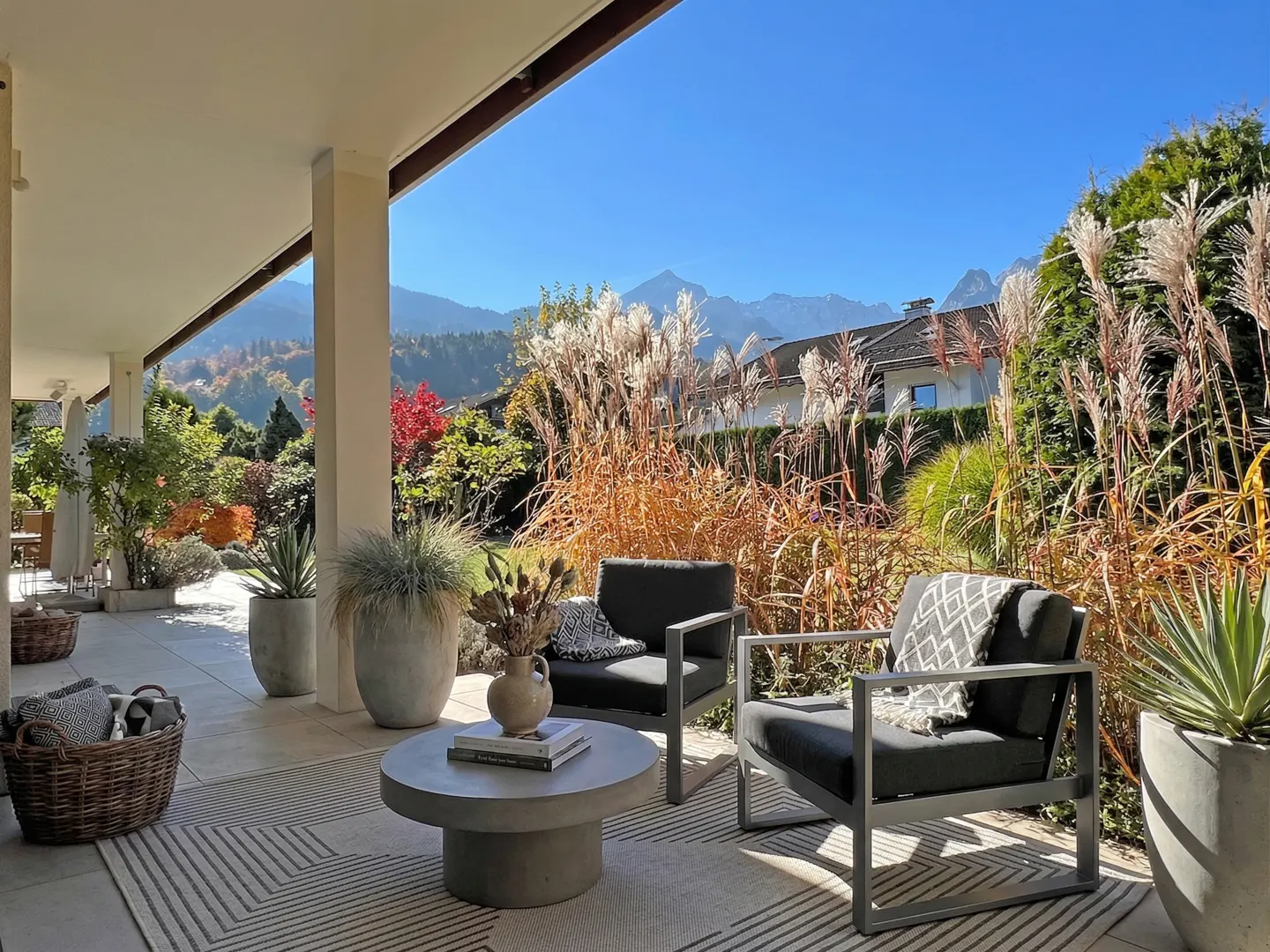 Outdoor patio with two gray chairs, a round table, and a patterned rug. Tall grasses and trees are in the background, with mountains in the distance.