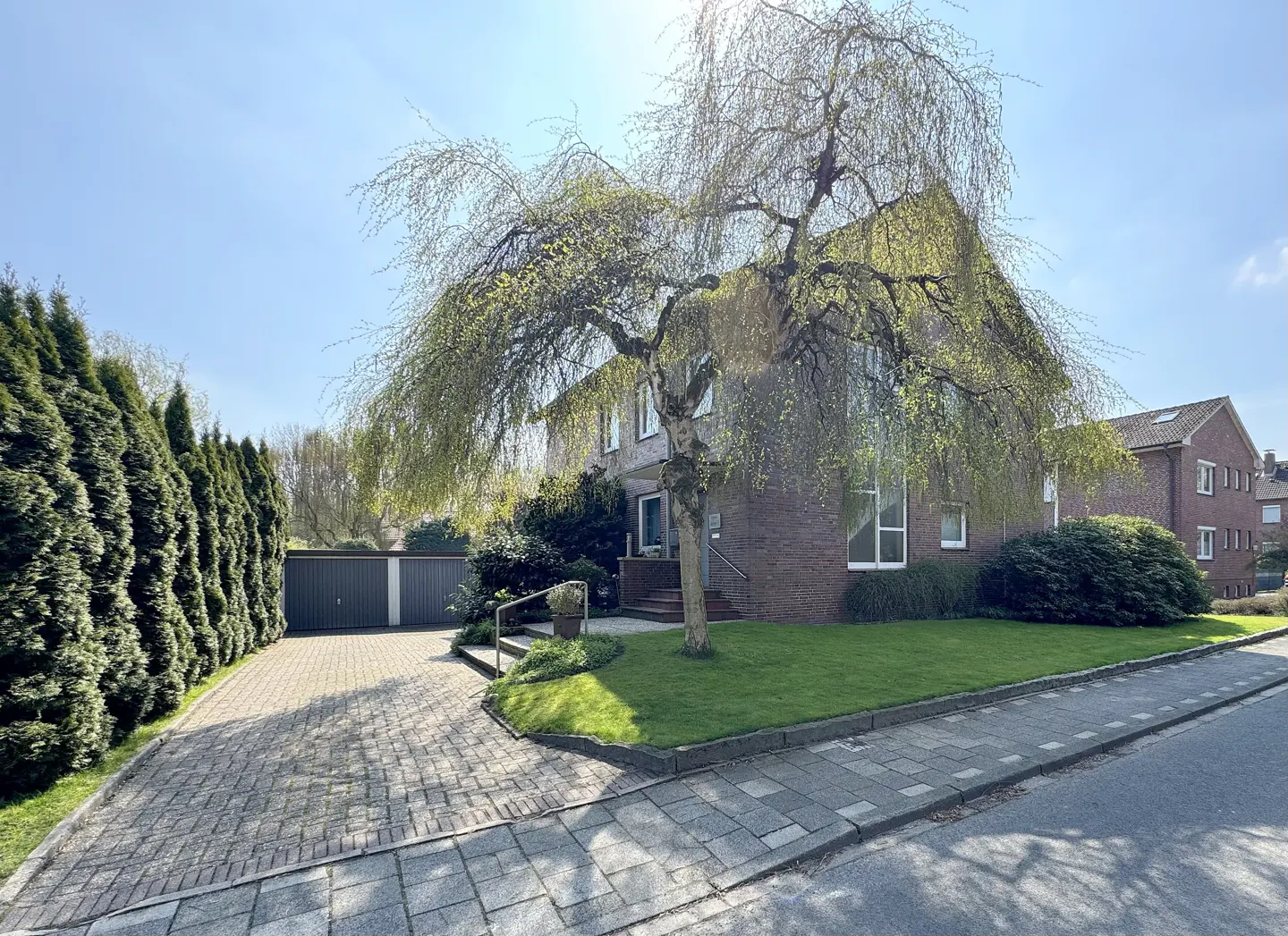 Brick house with a weeping willow tree in the front yard, a paved driveway, and a row of tall green bushes.