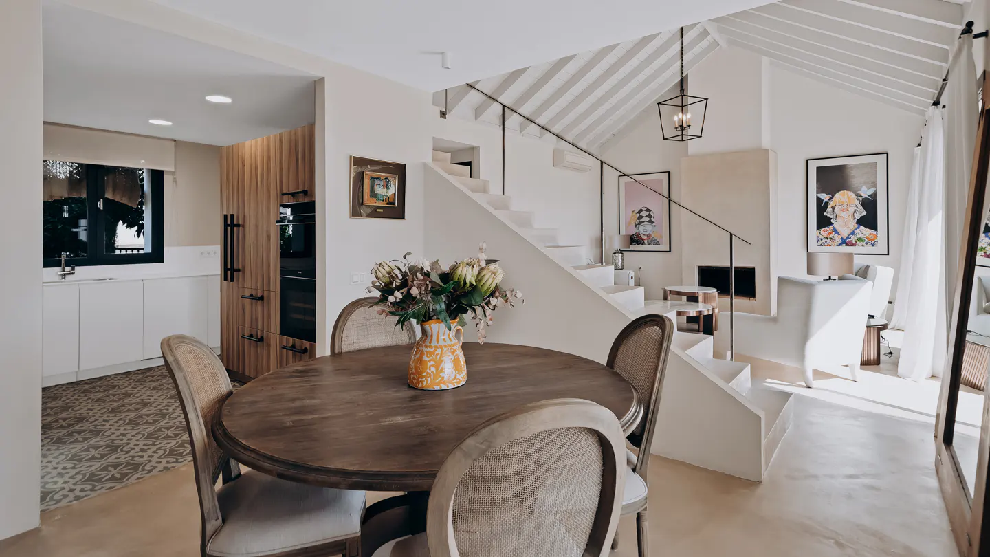 Bright, modern dining area with a round wood table, four chairs, and a vase of flowers. White staircase, kitchen, and artwork in the background.