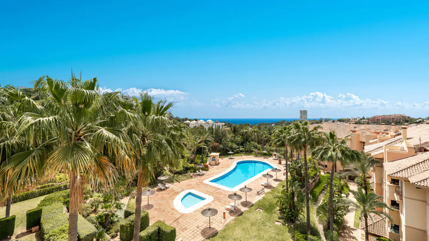 Aerial view of a resort with a blue pool, palm trees, and buildings under a clear blue sky.