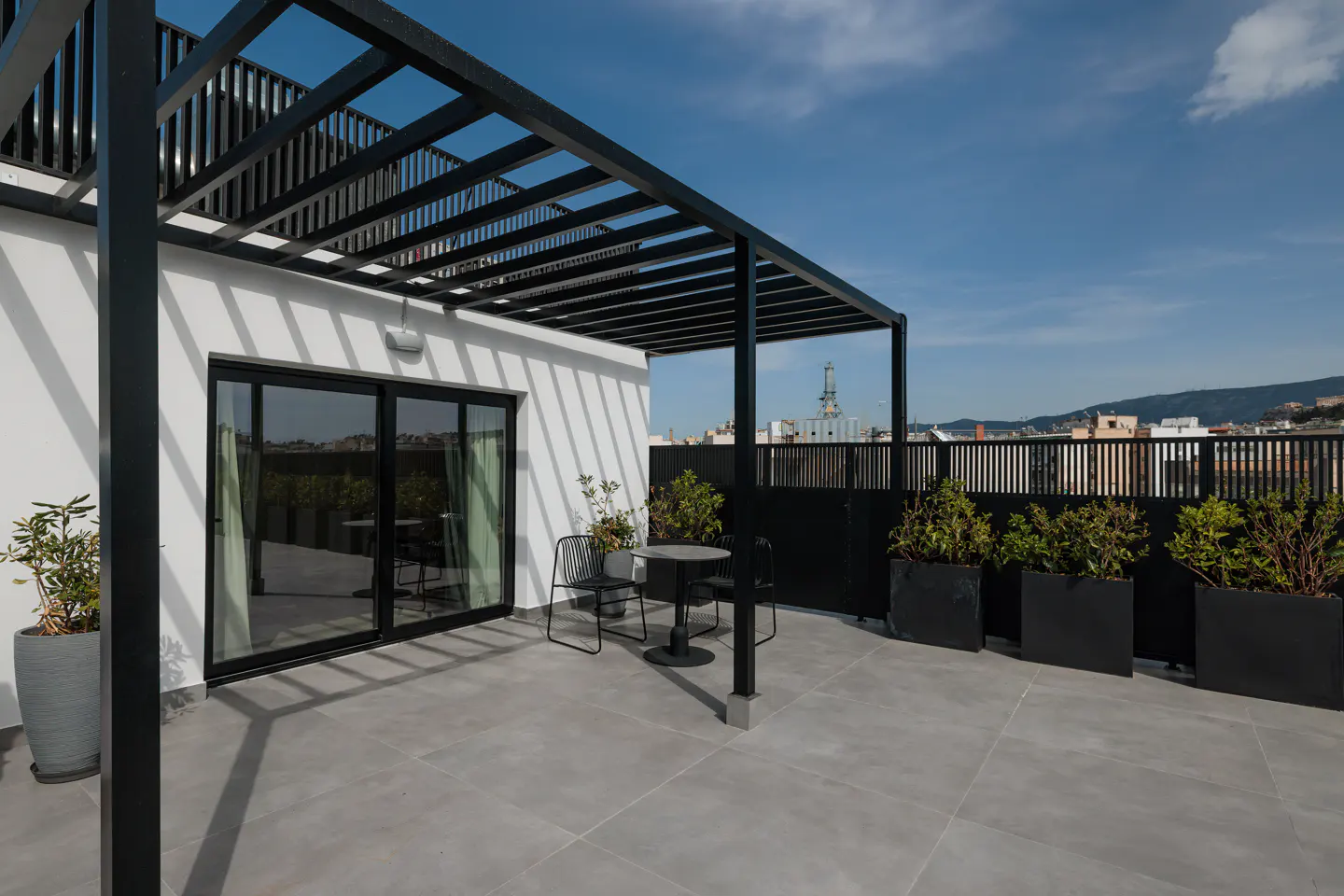 Rooftop patio with black pergola, table, and chairs. White building with sliding glass door. City skyline in the background under a blue sky.