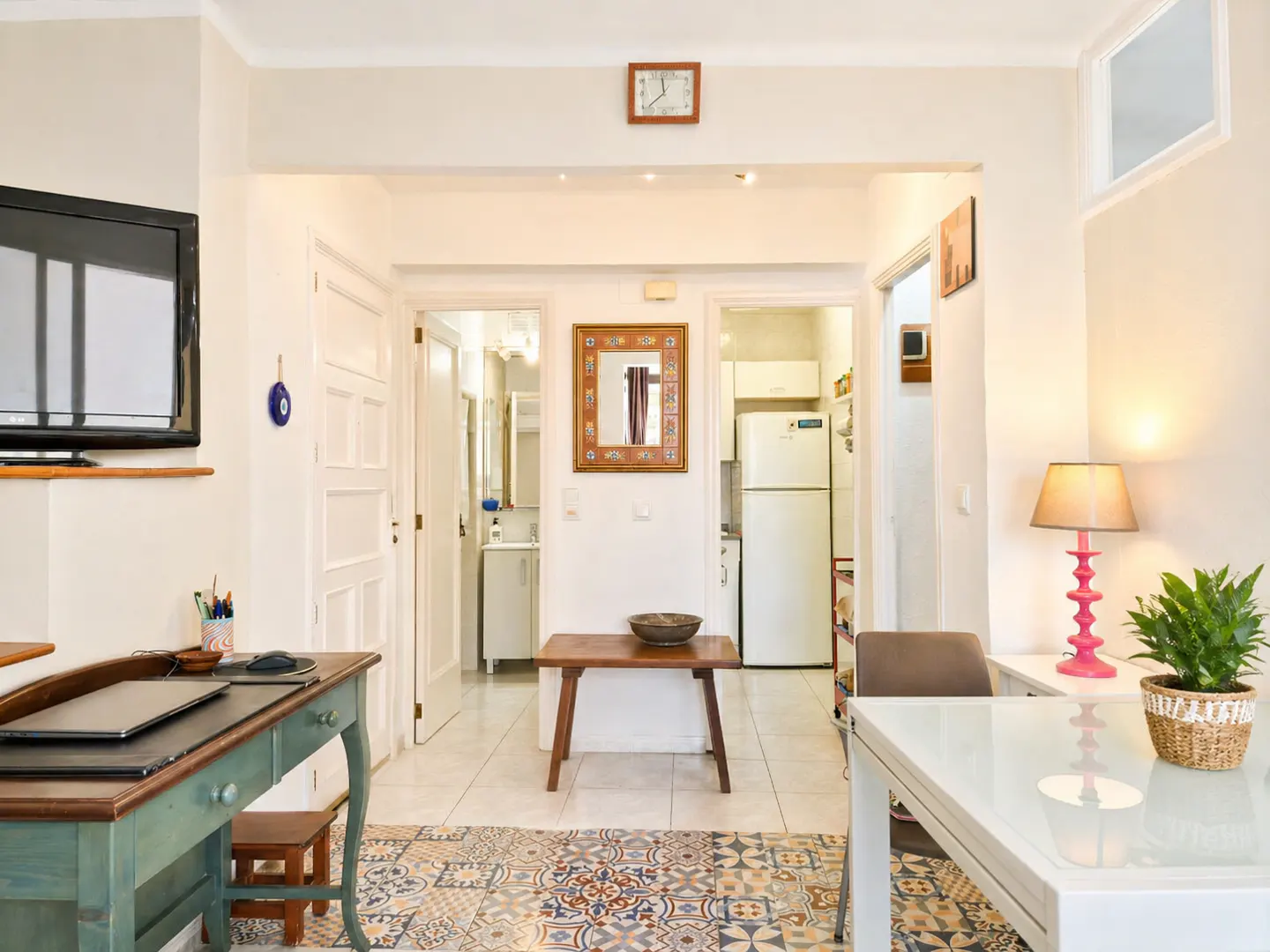 Bright apartment interior with a desk, table, and views into the bathroom and kitchen. The floor has colorful patterned tiles.