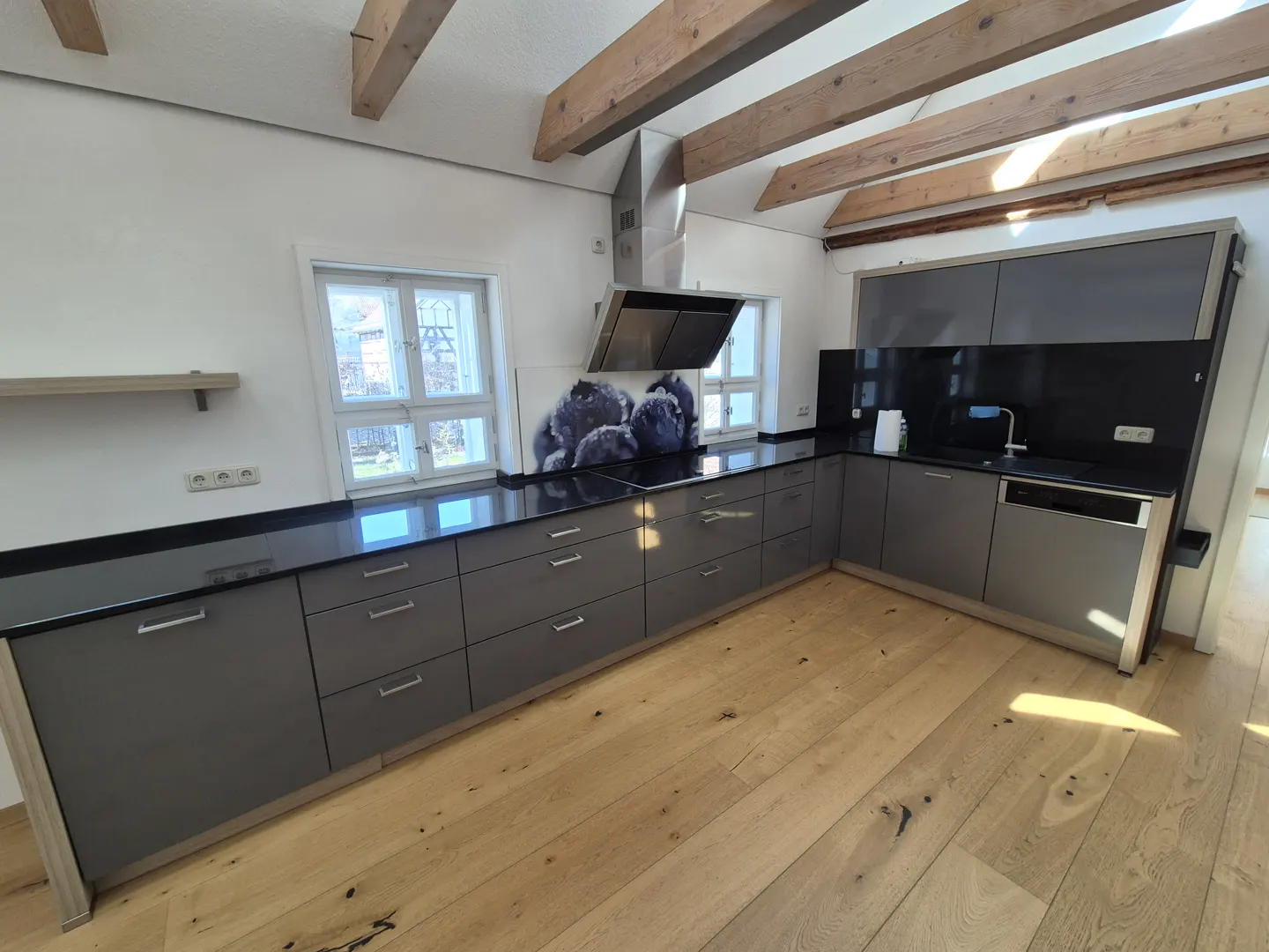 A modern kitchen with gray cabinets, black countertops, and light wood floors. Exposed wooden beams on the ceiling add rustic charm.