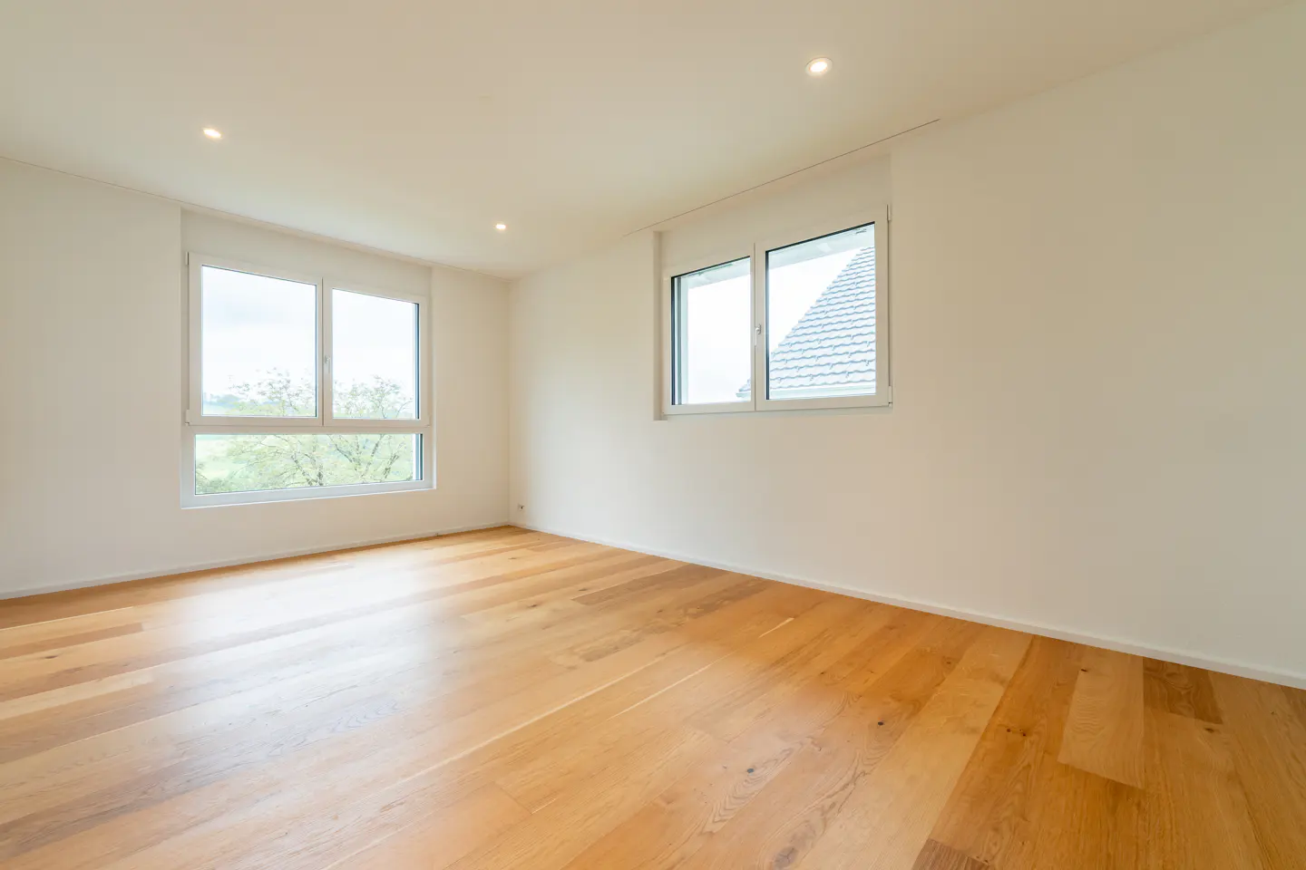 Empty room with white walls, light wood flooring, recessed ceiling lights, and two windows overlooking the outdoors.