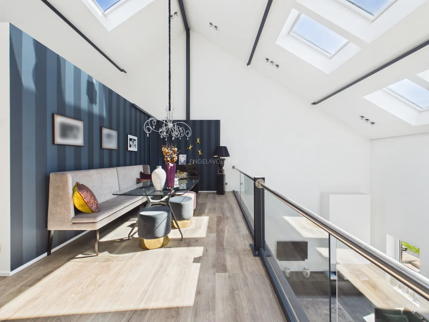 Interior view of a modern dining area with a long bench, glass table, and skylights. Blue striped wallpaper and a glass railing add to the contemporary design.