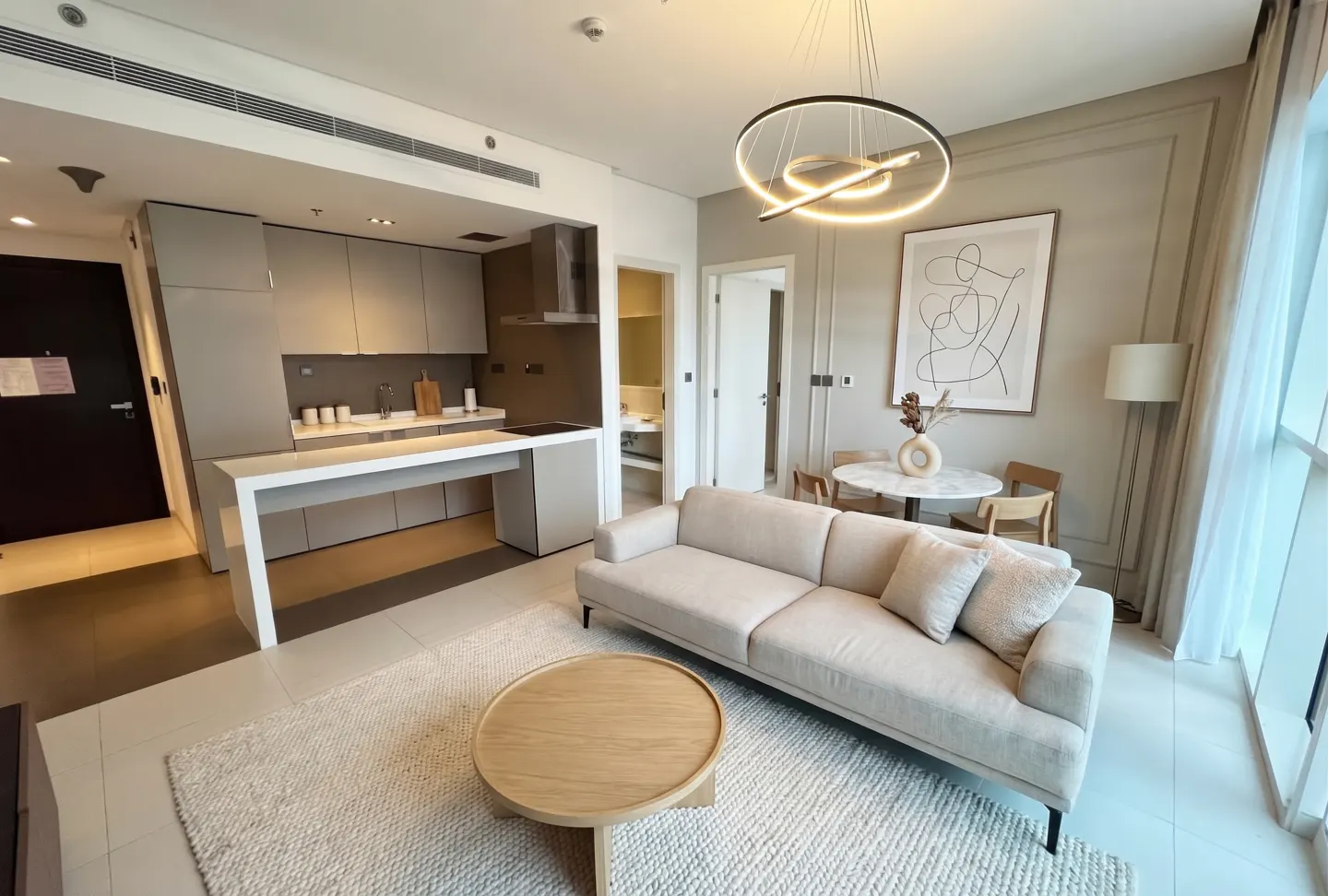 A modern apartment interior with a beige sofa, round wood table, and open kitchen. A circular light fixture hangs above.