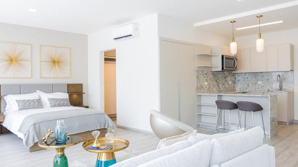 Bright studio apartment with a bed, kitchen, and living area. The walls are white, and the furniture is modern. There are two bar stools at the kitchen counter.