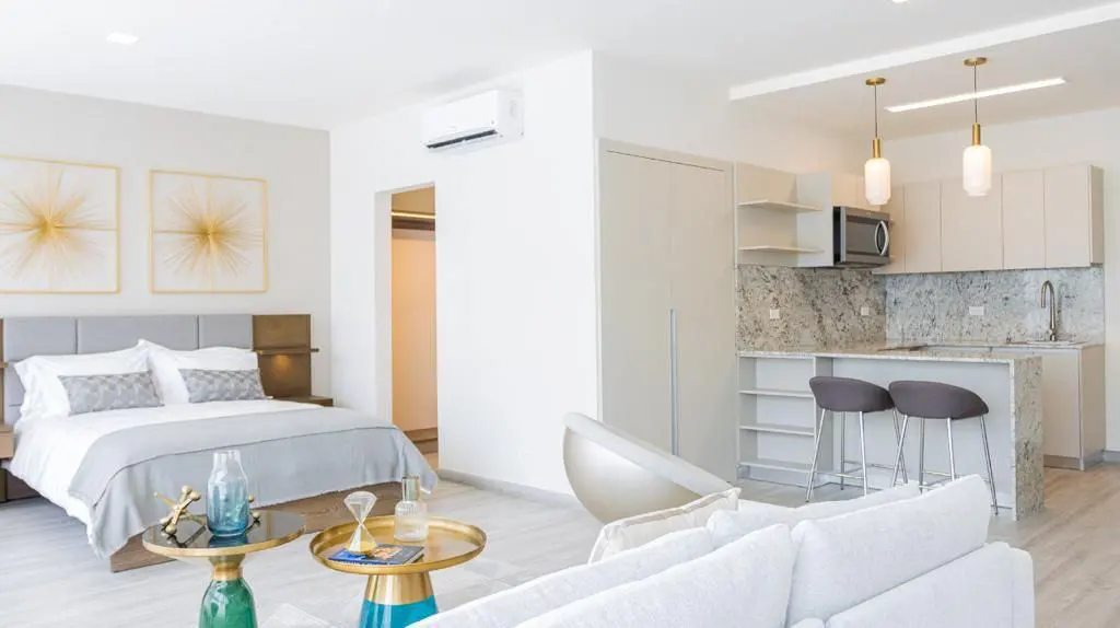 Bright studio apartment with a bed, kitchen, and living area. The walls are white, and the furniture is modern. There are two bar stools at the kitchen counter.