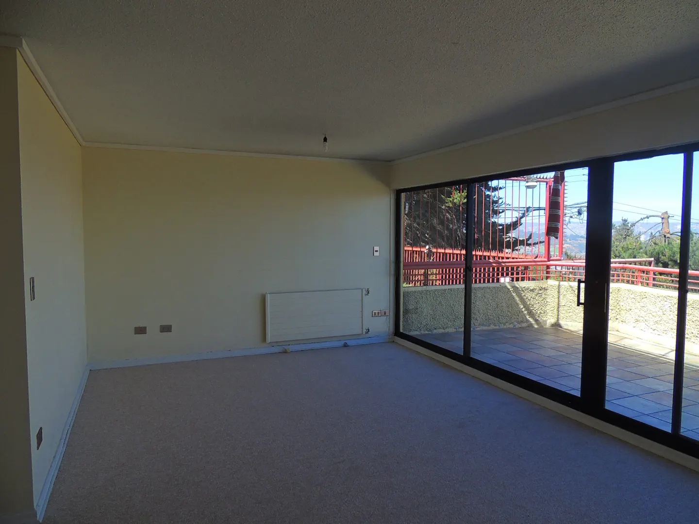 Bright, empty room with beige carpet and walls. Large sliding glass doors open to a patio with a red railing and a view of trees and mountains.
