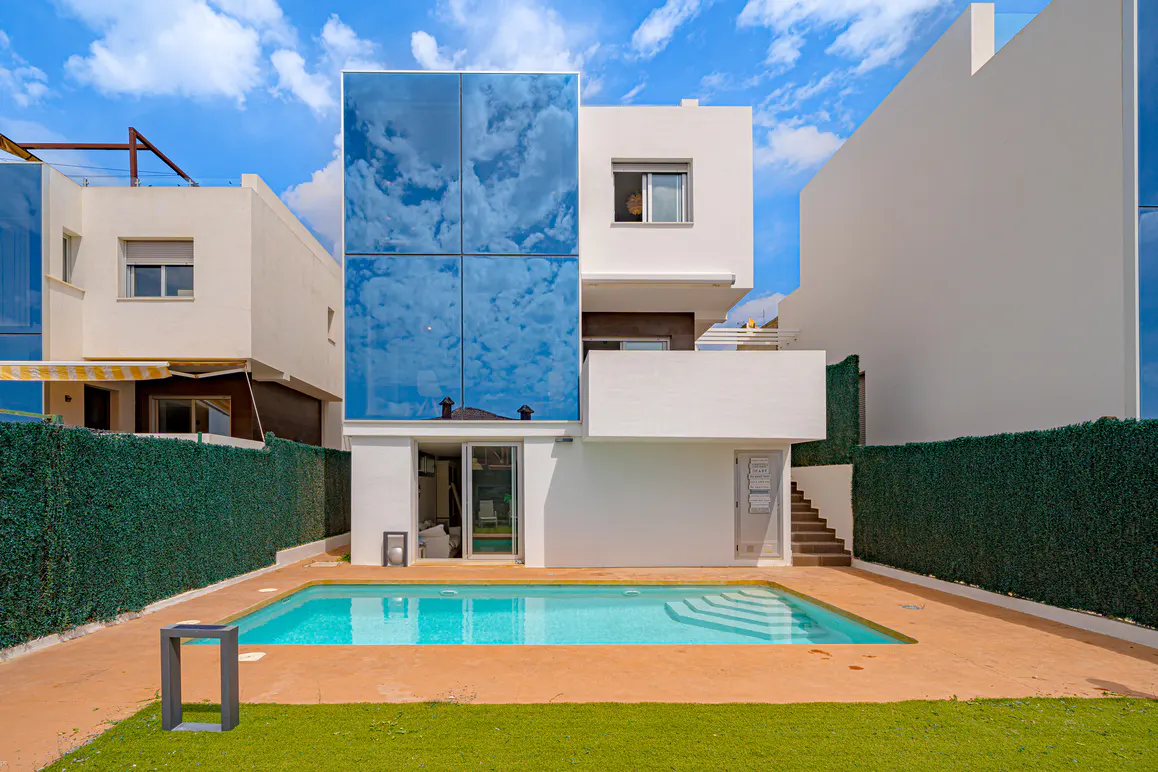 Modern white house with a pool. The house has a glass facade reflecting the blue sky and clouds. Green hedge surrounds the property.