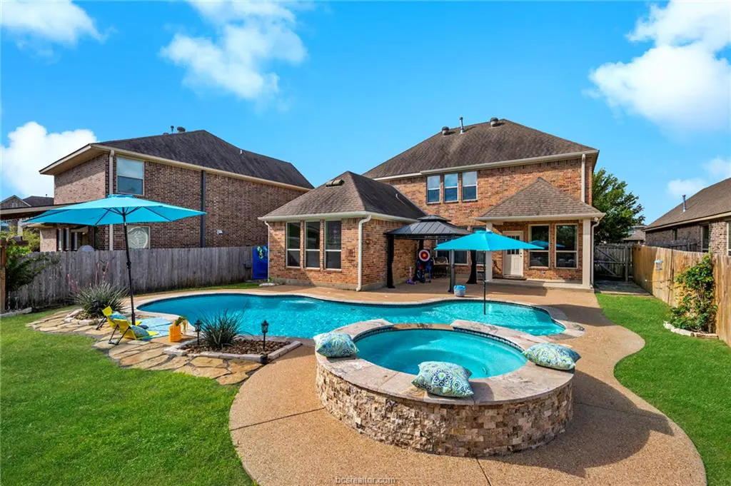Backyard view of a brick house with a pool, hot tub, and green lawn under a blue sky.