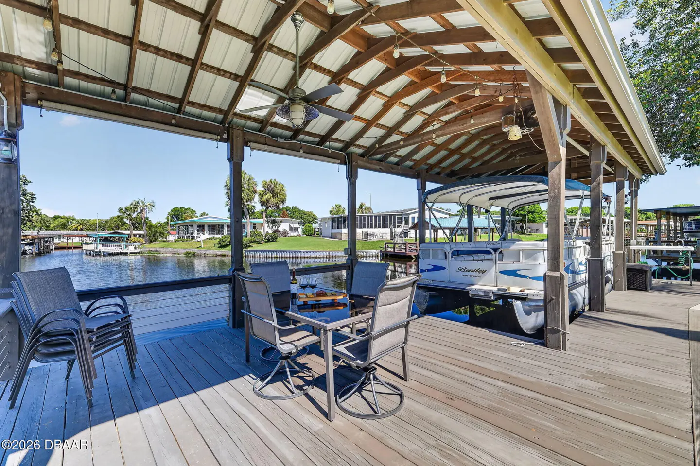 Covered boat dock with a table, chairs, and a boat. The dock is made of wood and has a metal roof. There is a ceiling fan and string lights.