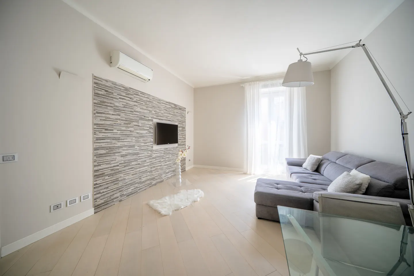 Bright living room with gray sectional sofa, glass table, stone accent wall with TV, and a tall silver floor lamp.
