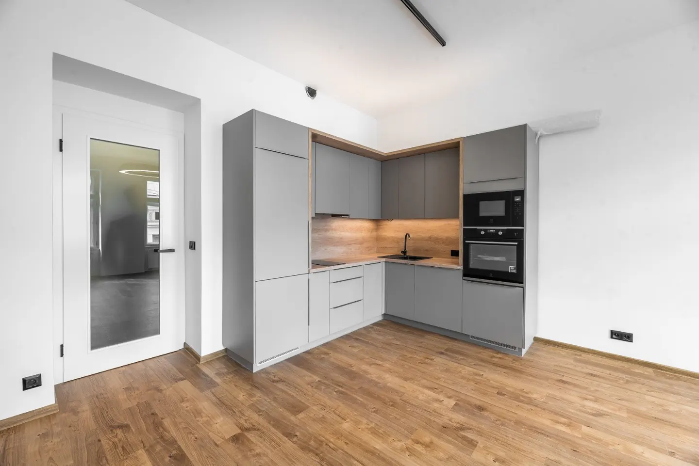 Bright, modern kitchen with gray cabinets, wood floors, and white walls. A glass door leads to another room. Black sink and appliances.