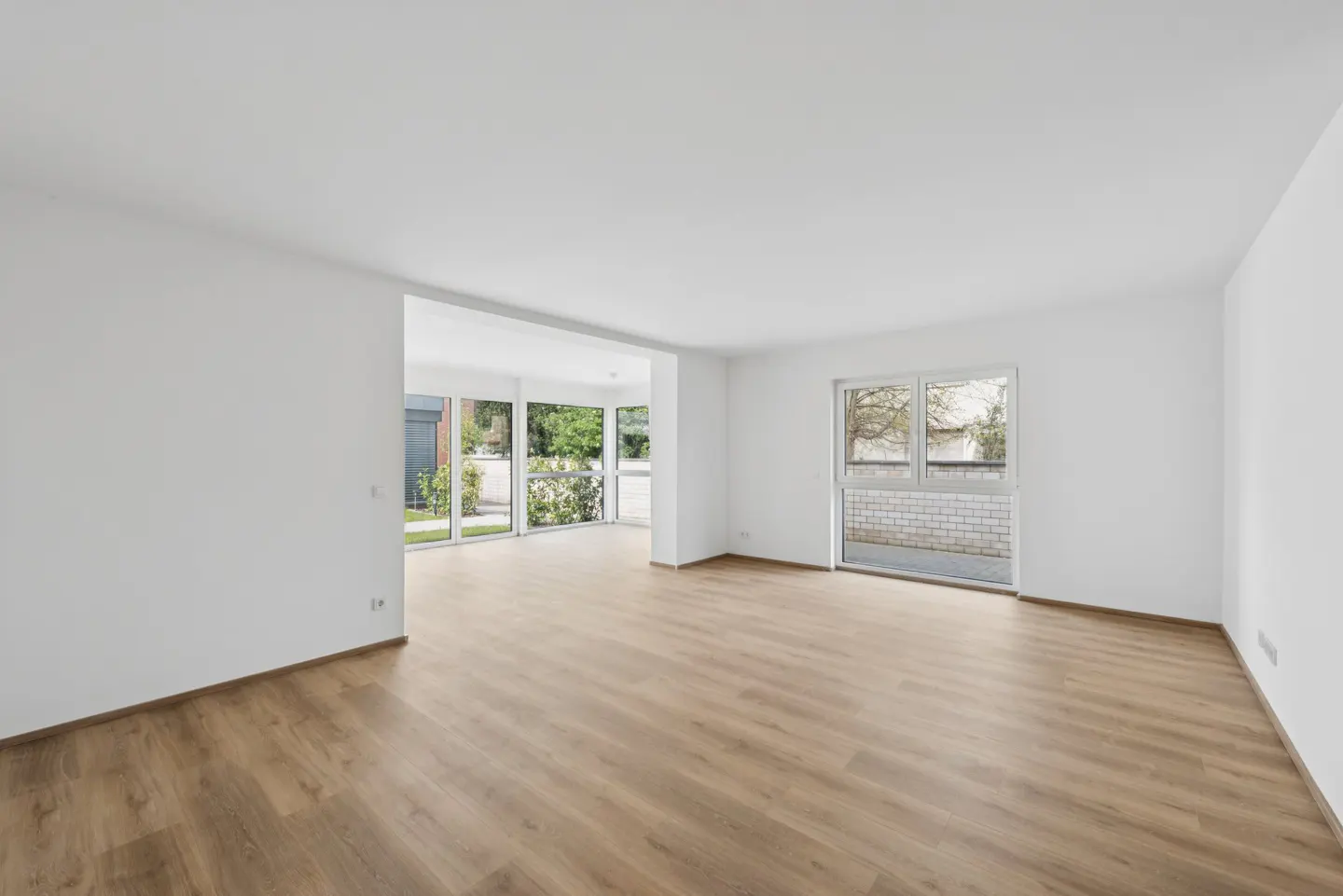 Bright, empty room with light wood floors and white walls. Large windows offer views of greenery and a brick wall.