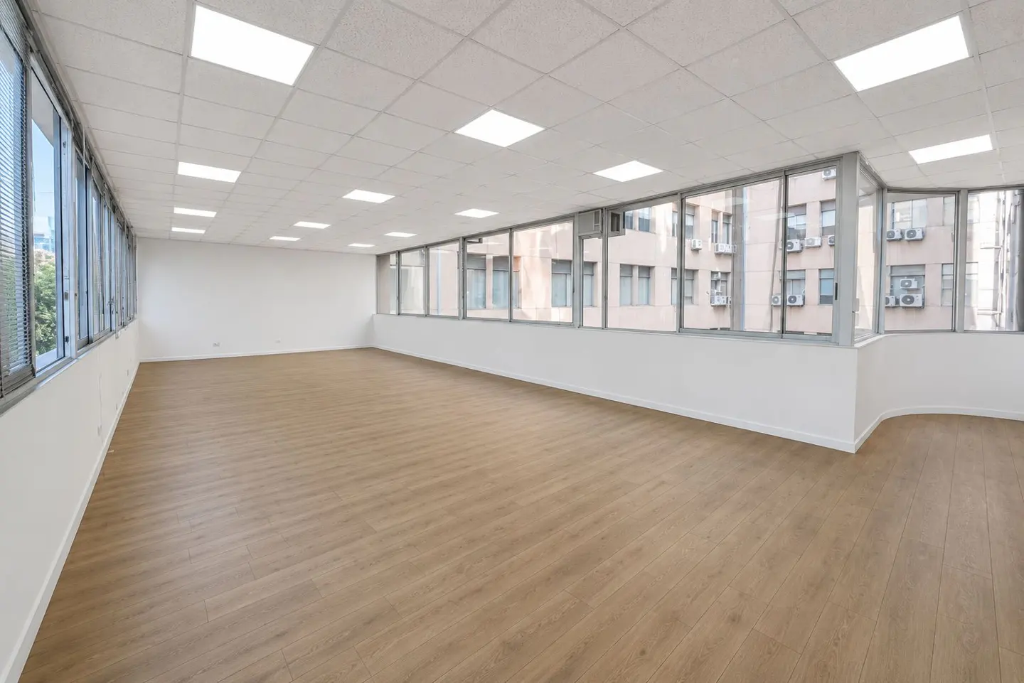 Bright, empty office space with wood floors, white walls, and large windows showing city buildings.