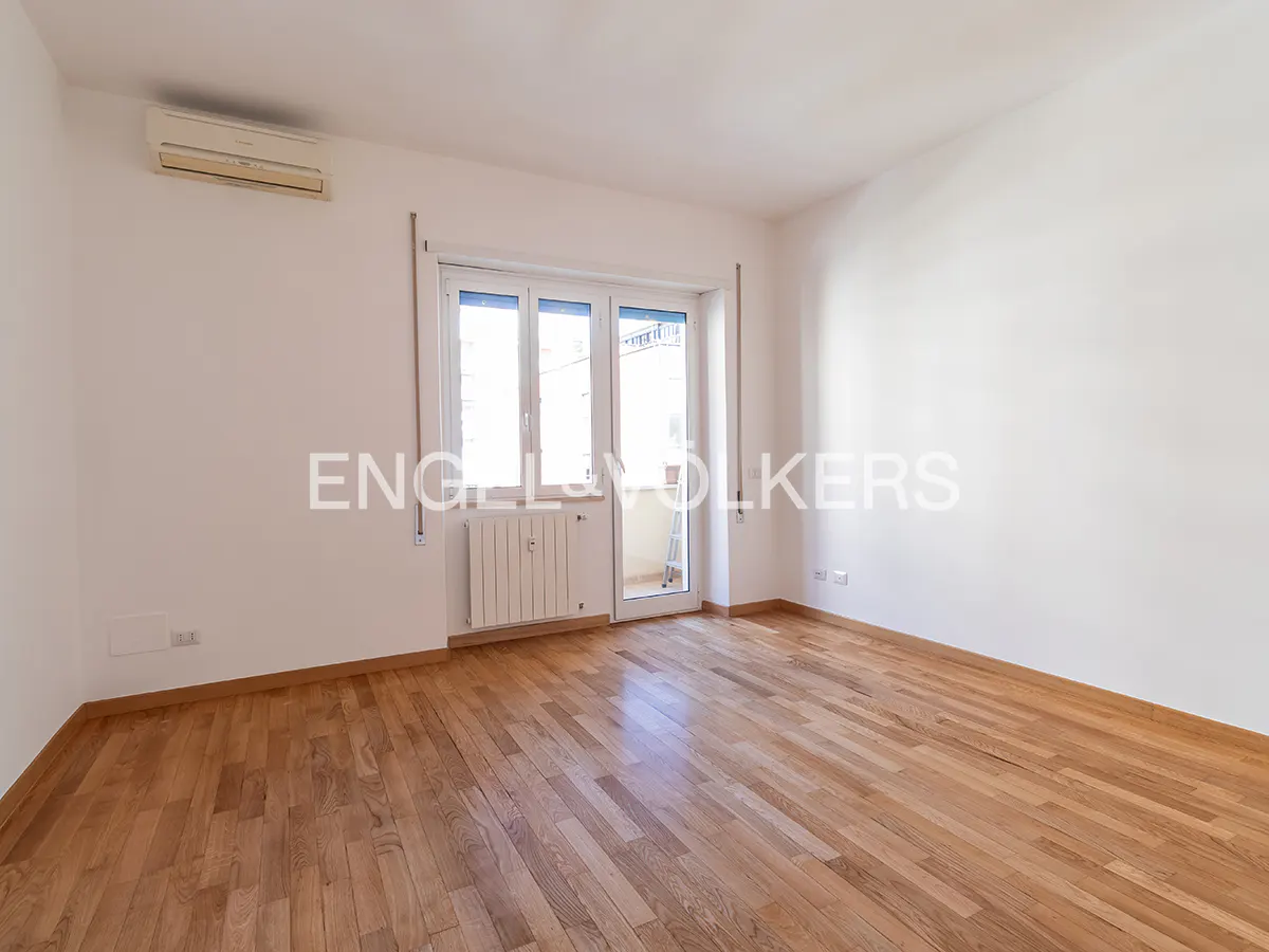 Empty room with hardwood floors, white walls, and a balcony door. A radiator sits below the door, and an AC unit is mounted high on the wall.