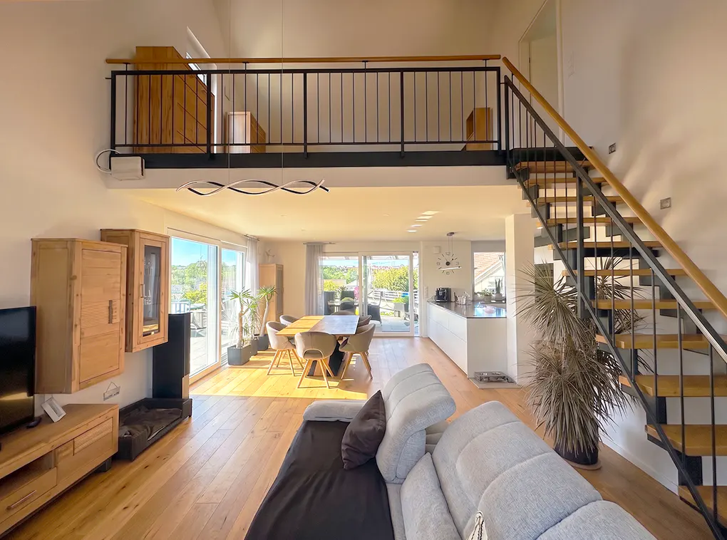 Open-plan living room with light wood floors, a grey sofa, dining area, modern kitchen, and a loft with a metal staircase.
