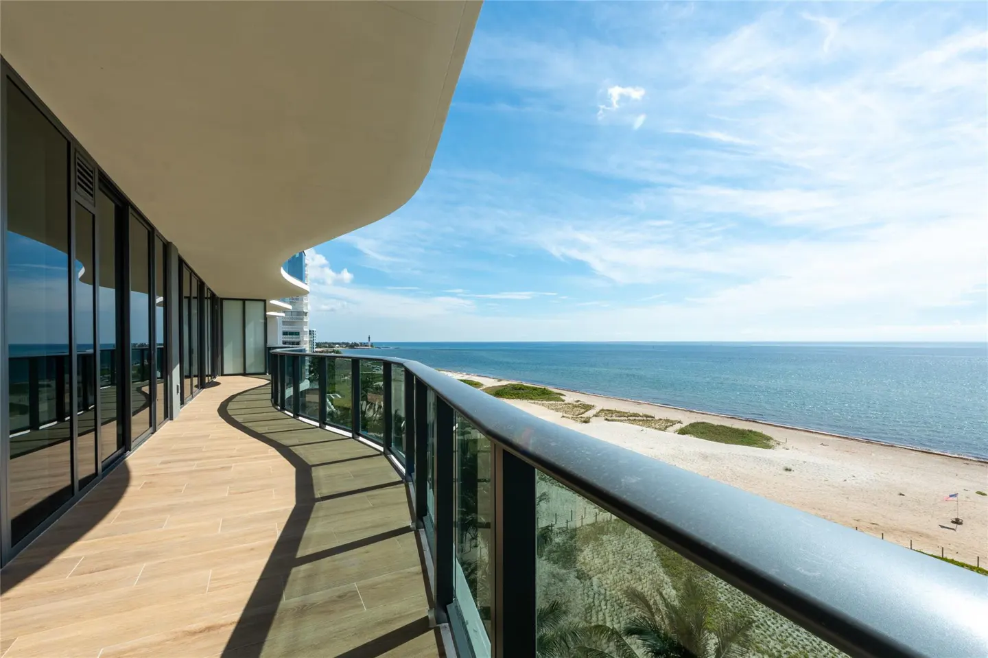 Curved balcony with glass railings overlooking a beach and ocean on a sunny day.