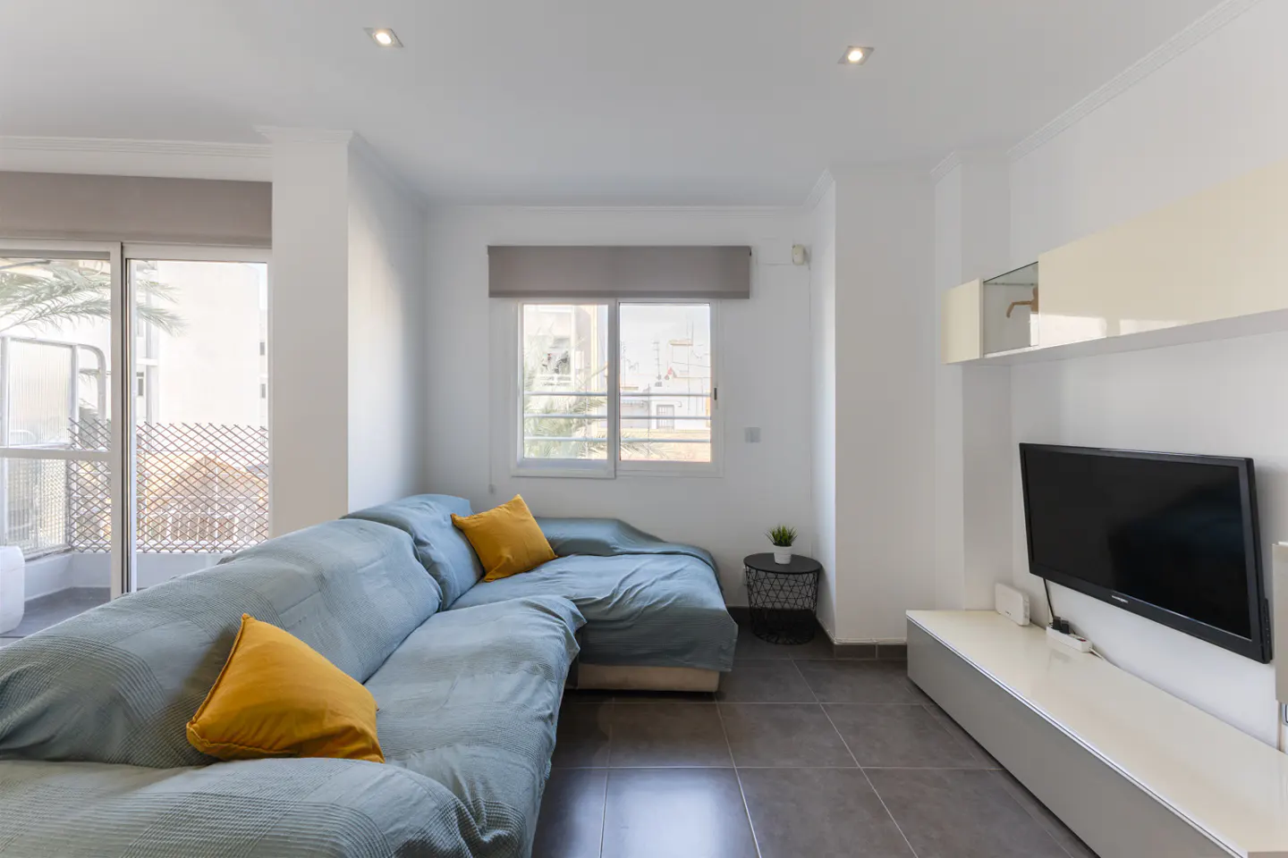 Bright living room with a large blue sectional sofa, yellow pillows, a TV, and a balcony.