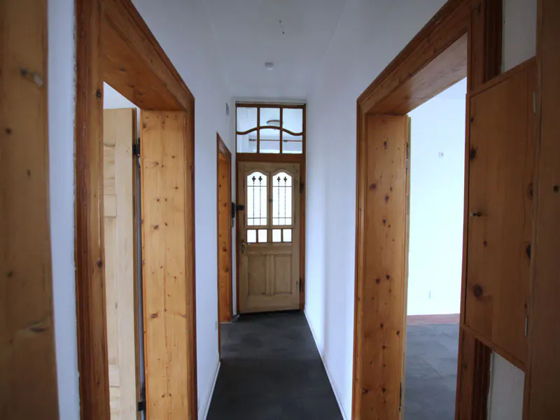 Hallway with white walls, dark floor, and wooden doors. A front door with glass panes is at the end of the hall.