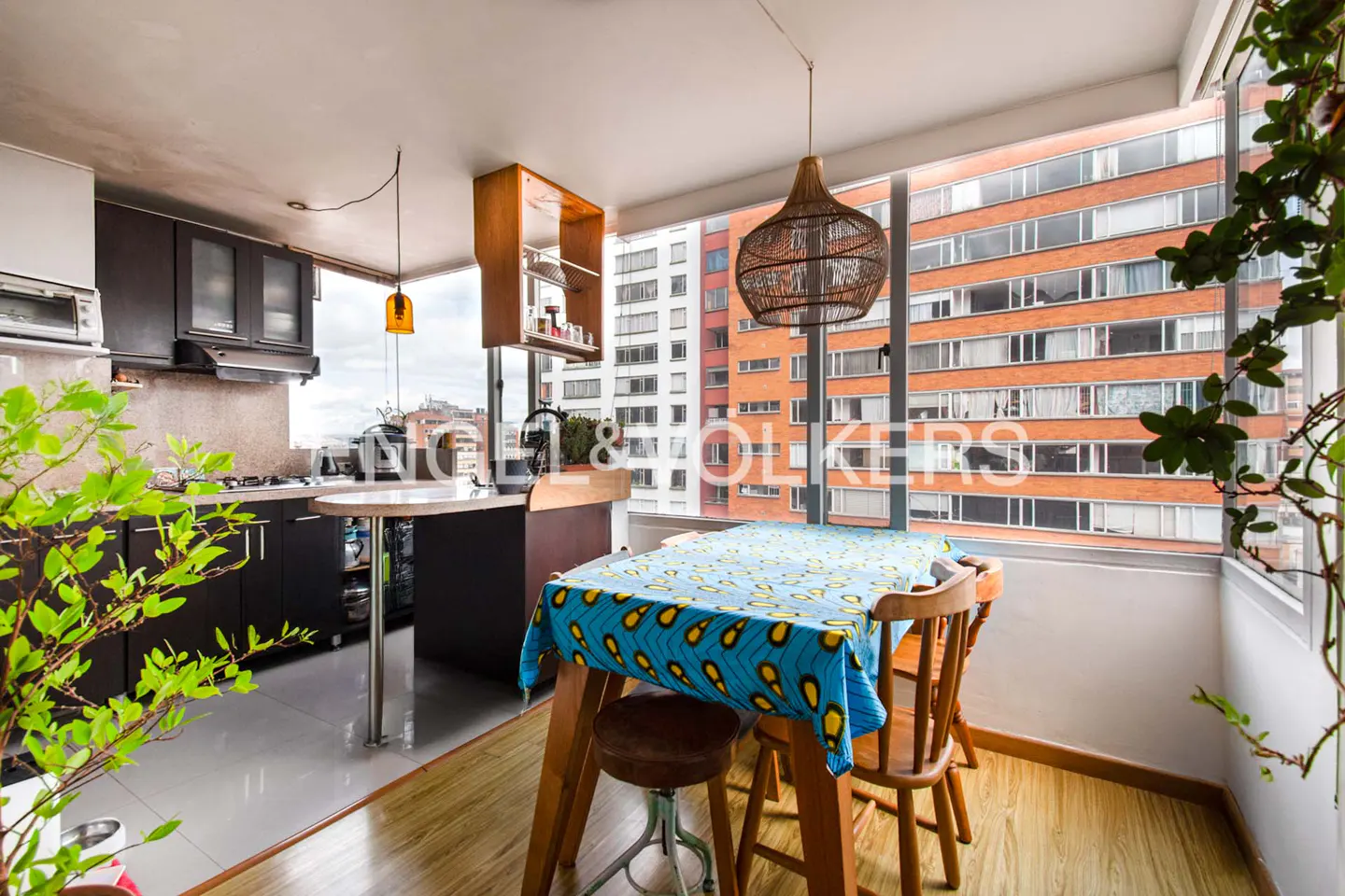 A bright kitchen with a dining table covered in a blue patterned cloth, wooden chairs, and a view of city buildings.