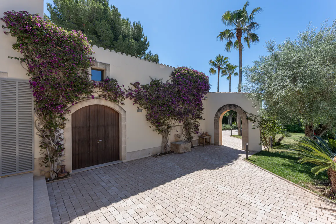 A paved courtyard with a wooden arched door, stone archway, and walls covered in vibrant purple bougainvillea under palms.
