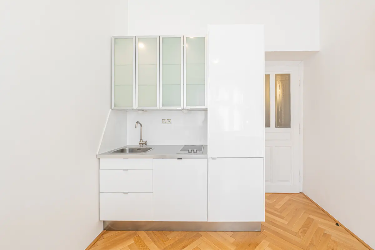 A small, modern, all-white kitchen with stainless steel sink and light wood herringbone floor. A white door is visible in the background.