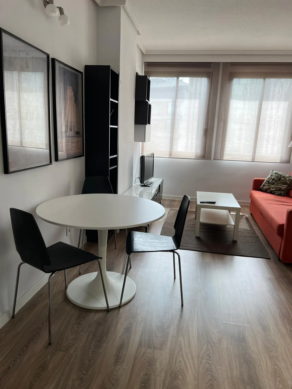 A bright living room with a white table, black chairs, an orange sofa, and wood floors. Two framed pictures hang on the wall.