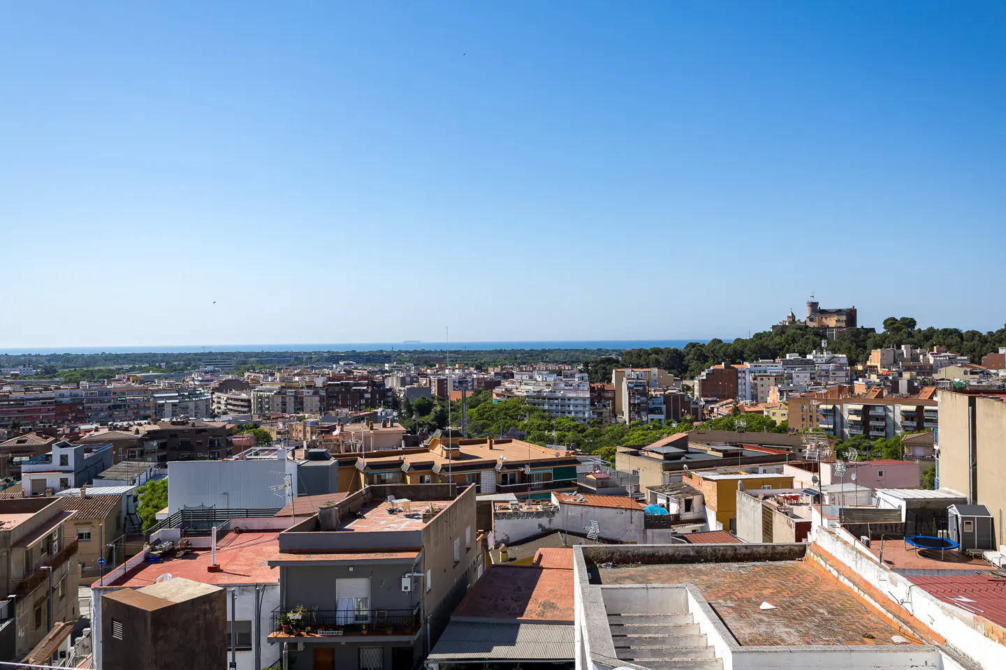 Elevated view of a Mediterranean town with flat-roofed buildings, lush trees, and a distant castle overlooking the sea.