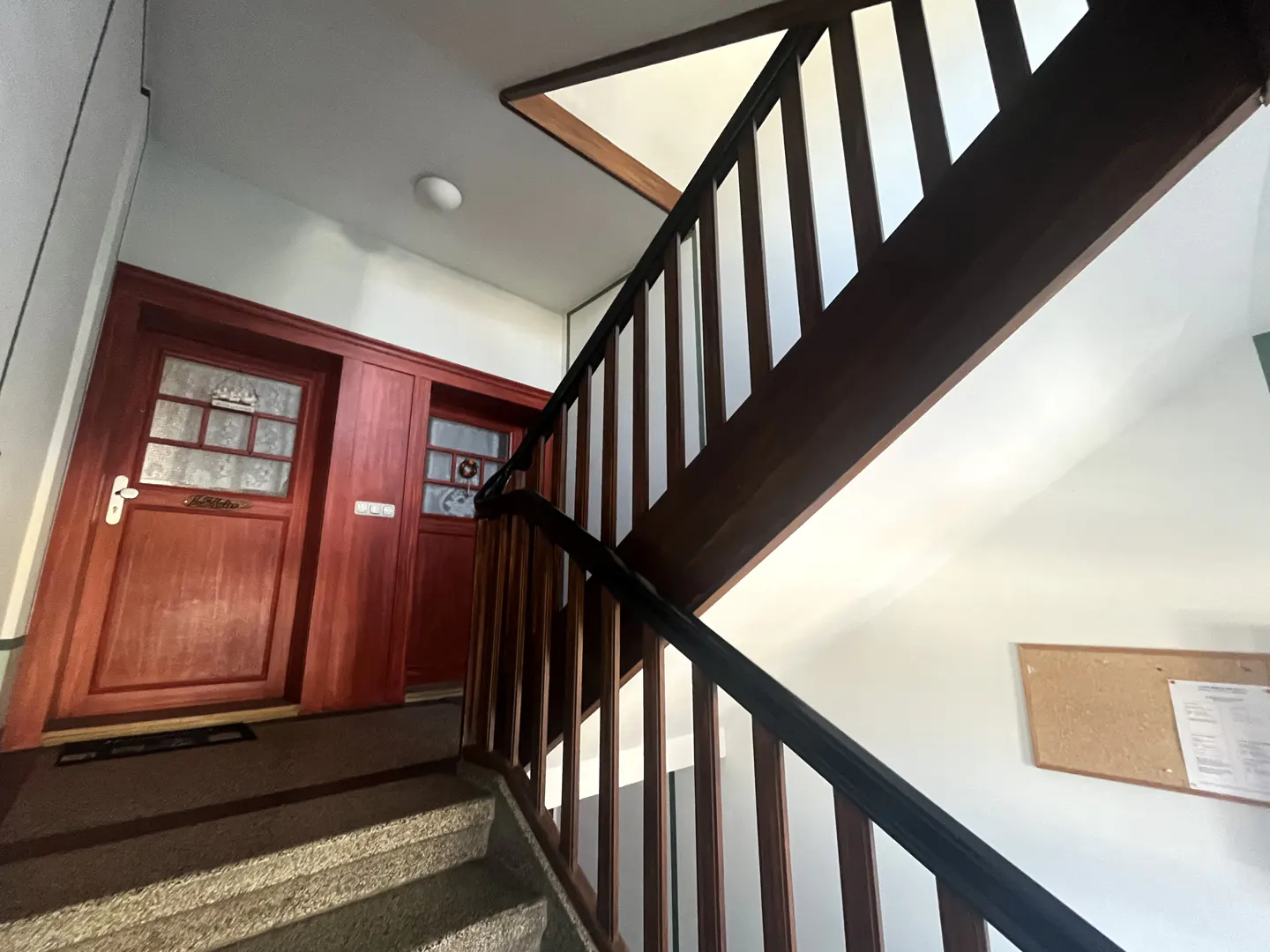 Interior view of a staircase with brown wooden railings, leading to two red doors with glass panels. A corkboard hangs on the white wall.