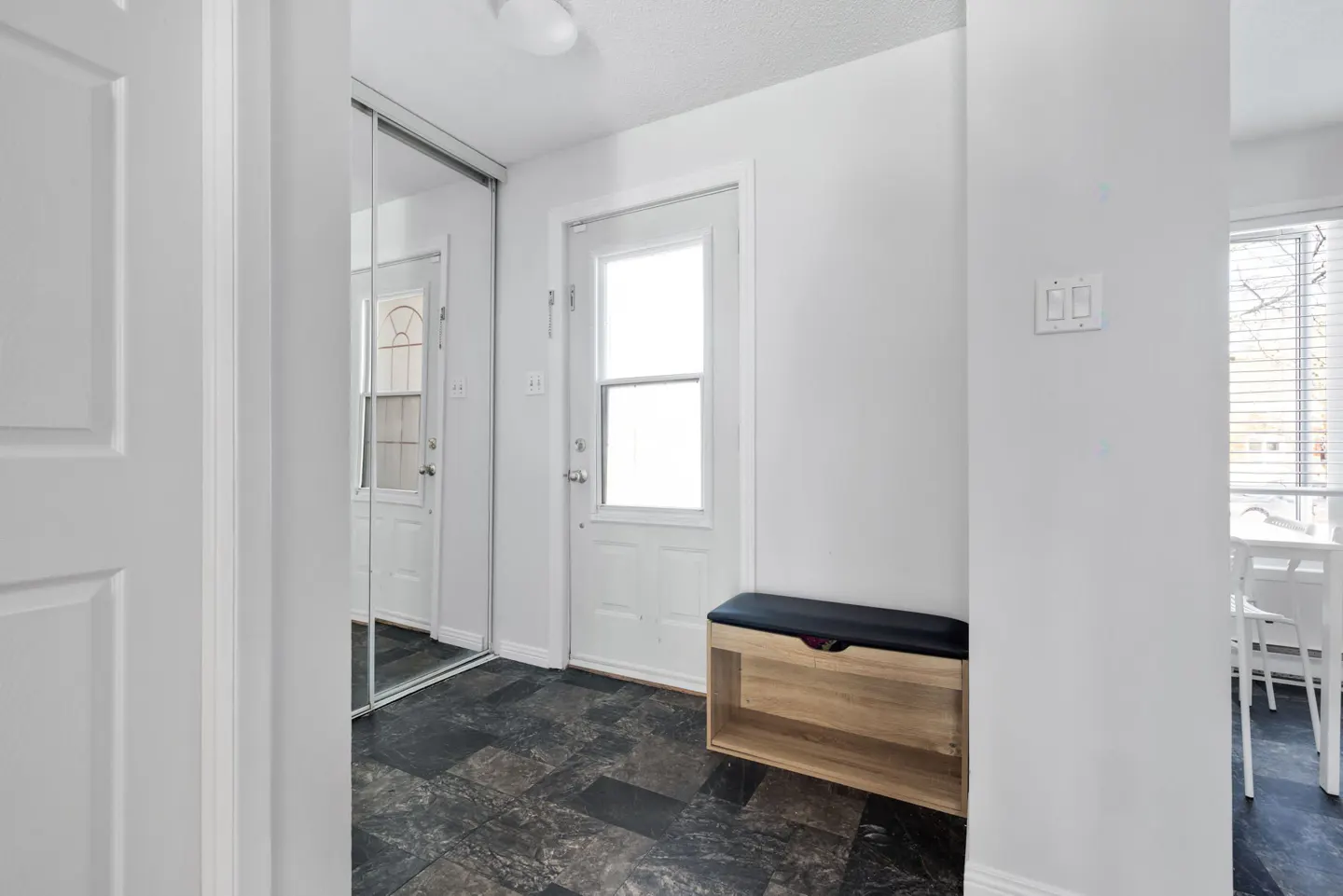 Entryway with dark tile floor, white walls, mirrored closet, and a wooden bench with a black cushion. A white door leads outside.
