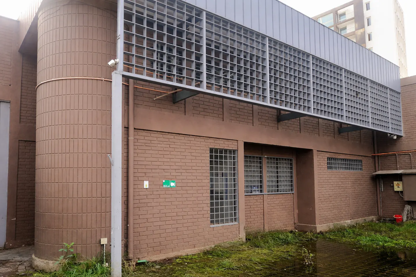 Exterior view of a brown brick building with metal grates above windows and a cylindrical section. Mossy ground and a red bucket are visible.