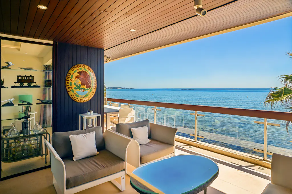 Outdoor patio with ocean view. Two chairs with white pillows, blue table, and glass railing. Mexican coat of arms on the wall.