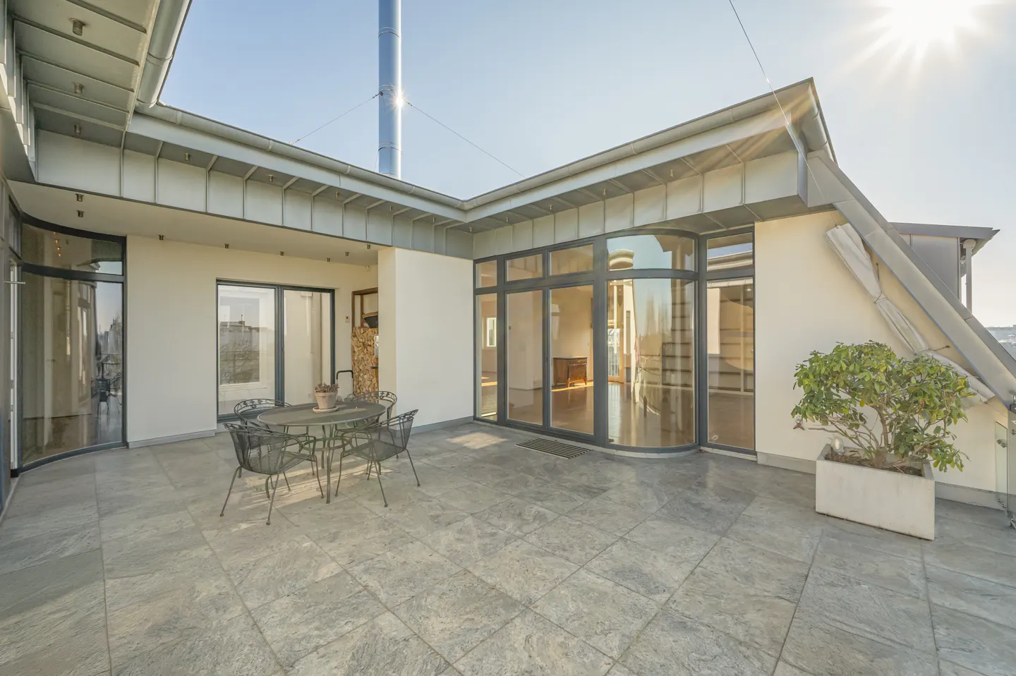 Outdoor patio with gray stone tiles, a metal table and chairs, and a potted plant. Large windows and a metal chimney are visible.