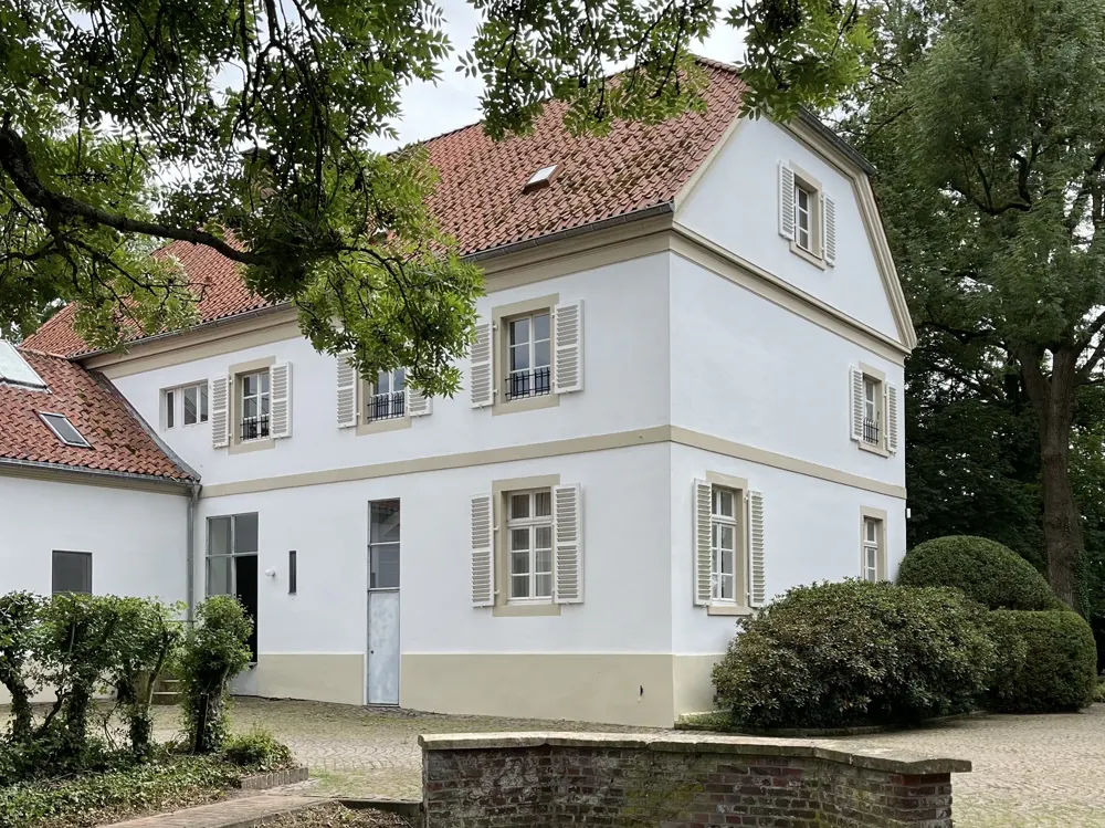 Two-story white house with a red tile roof and white shutters, surrounded by green trees and bushes.