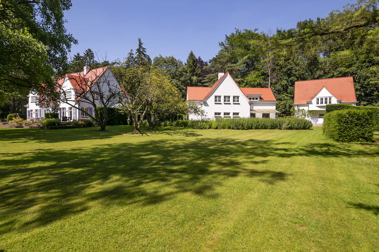 Three white houses with red roofs sit on a large green lawn surrounded by trees under a blue sky.