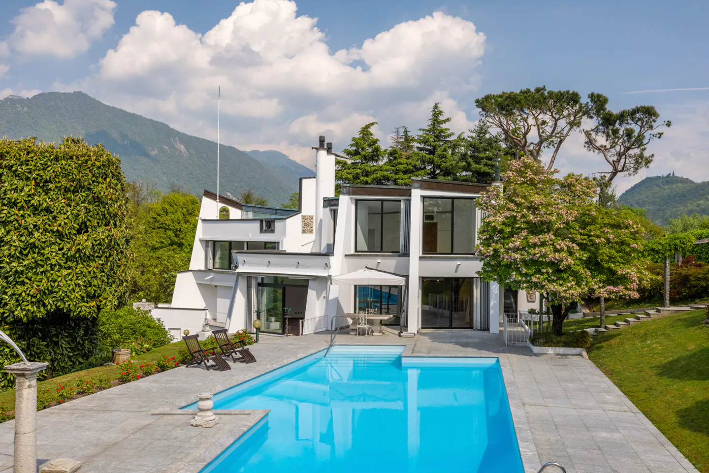 Modern white house with a blue pool, stone patio, and green trees. Mountains are visible in the background under a partly cloudy sky.