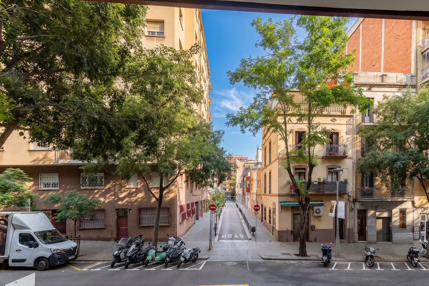 View from above of a narrow city street lined with tall apartment buildings, green trees, parked scooters, and a white van.