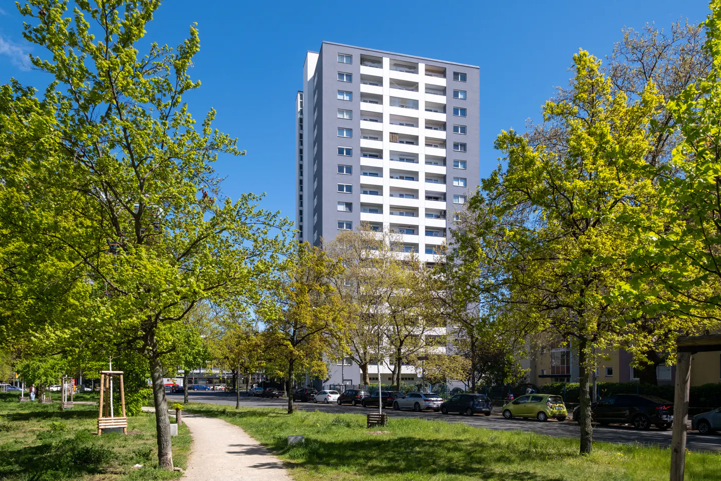 Tall gray and white apartment building with balconies, framed by green trees under a clear blue sky. A path and street with parked cars are in the foreground.