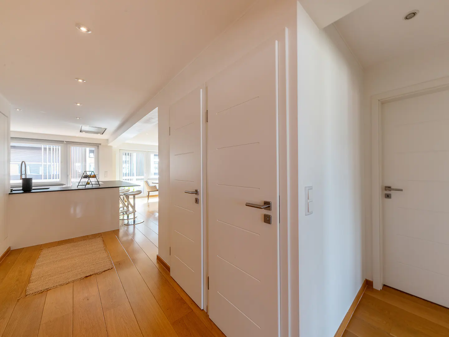 Bright, modern apartment hallway with wood floors, white walls, and doors. Kitchen island visible in the background.