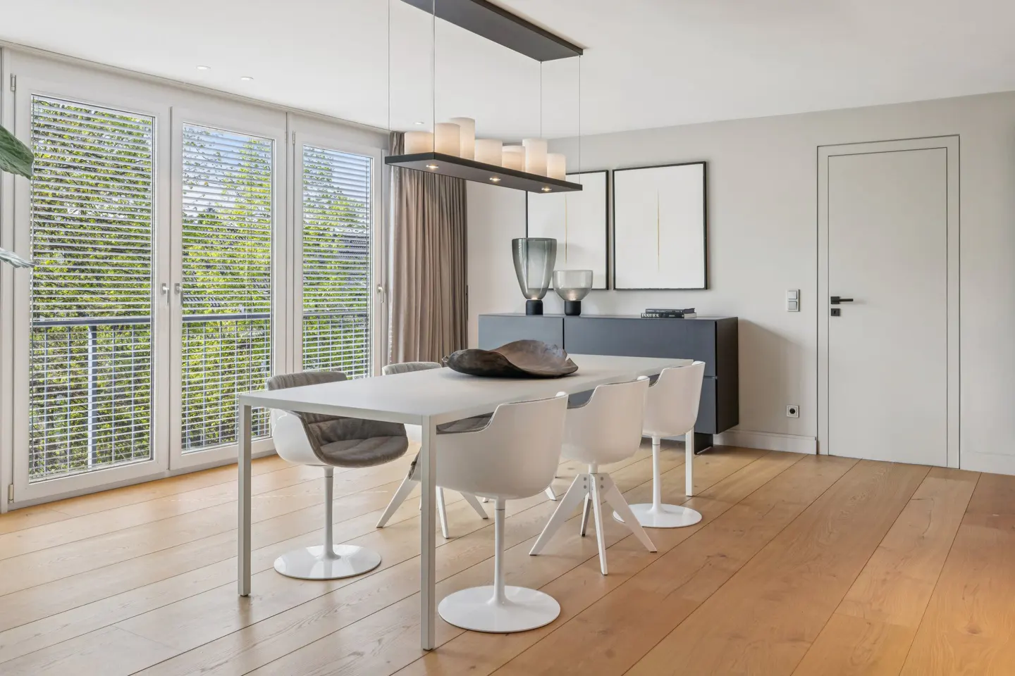 Bright dining room with wood floors, a white table and chairs, and a modern light fixture. Large windows with blinds let in natural light.