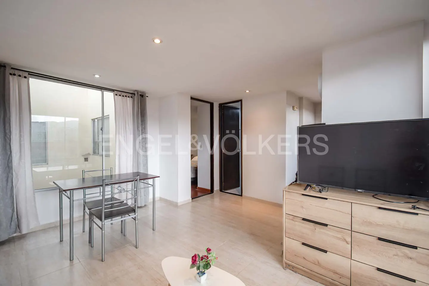 Bright living area with a light wood dresser, flat-screen TV, small dining table, and a view of an outdoor patio.