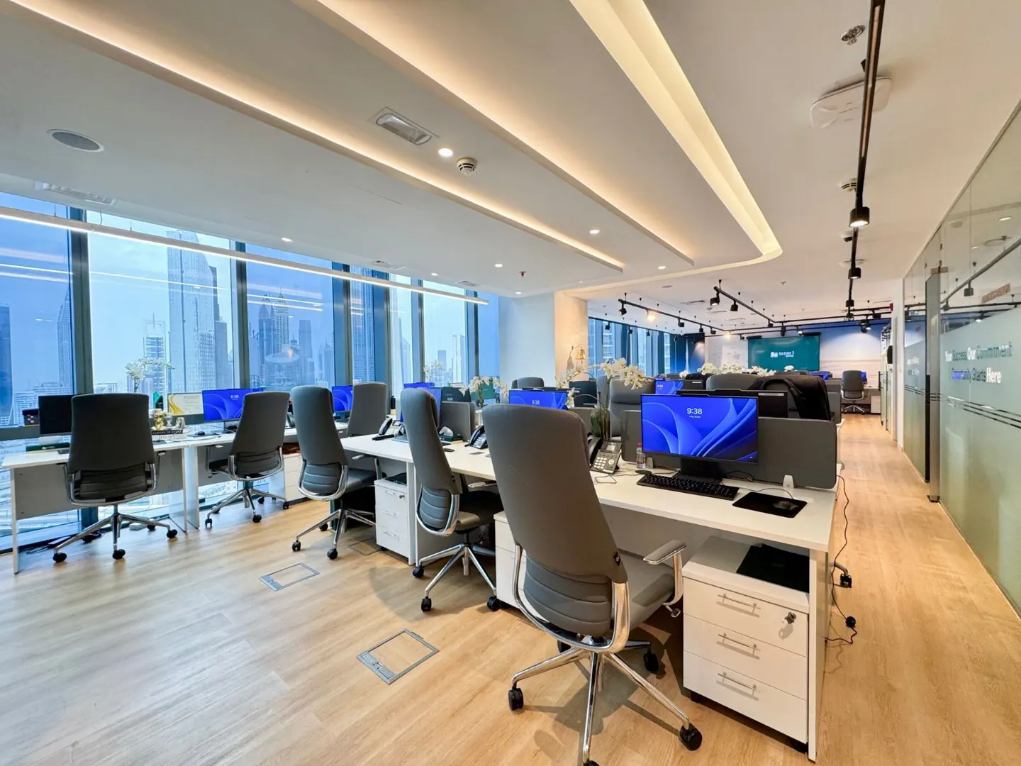 Bright, modern office space with white desks, gray chairs, and large windows overlooking a city skyline. Computers are on each desk.