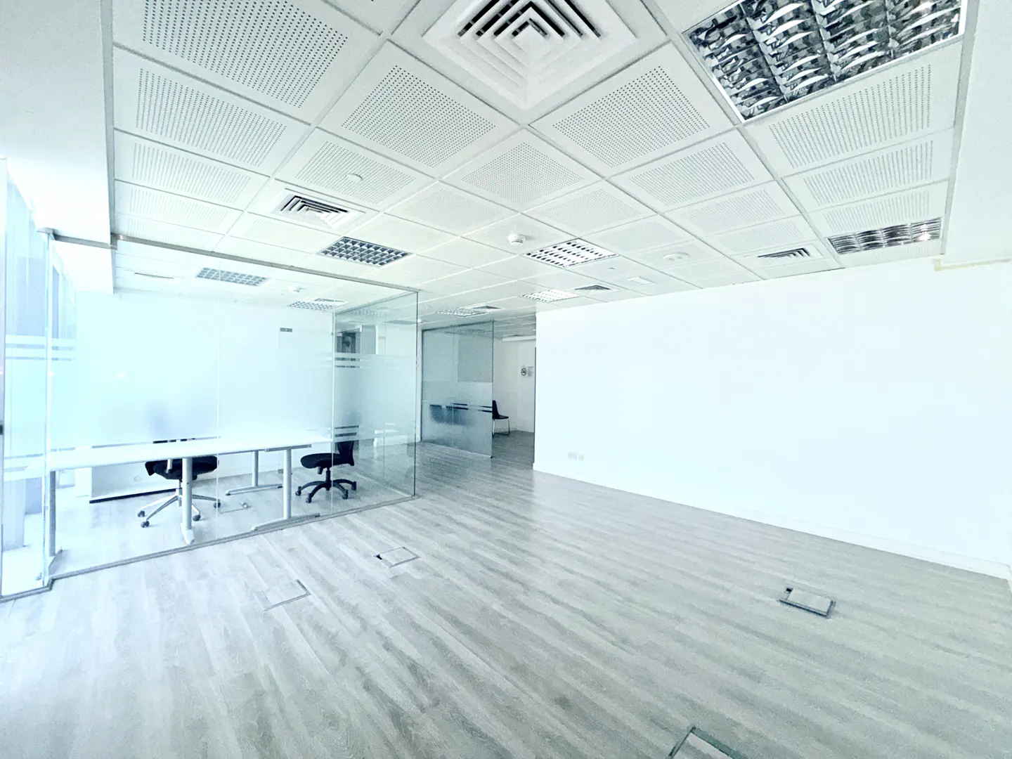 Bright, empty office space with glass-walled rooms, white walls, and a gray wood floor. The ceiling has square panels and recessed lighting.