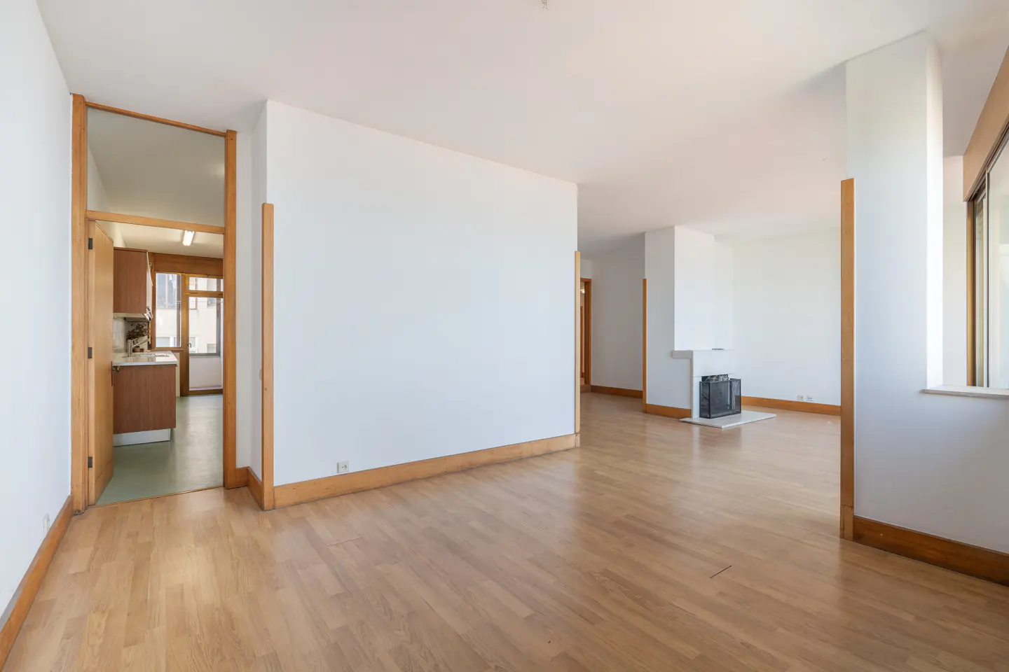 Bright, empty room with wood floors, white walls, and wood trim. A kitchen is visible through a doorway. A fireplace is in the background.