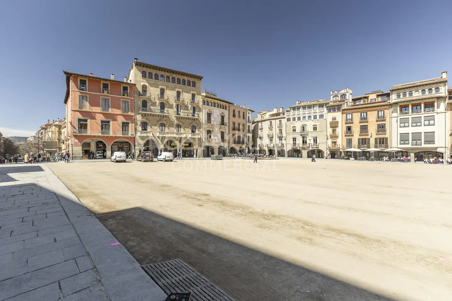 Wide shot of a town square with beige sand and buildings with arches and balconies under a clear blue sky.