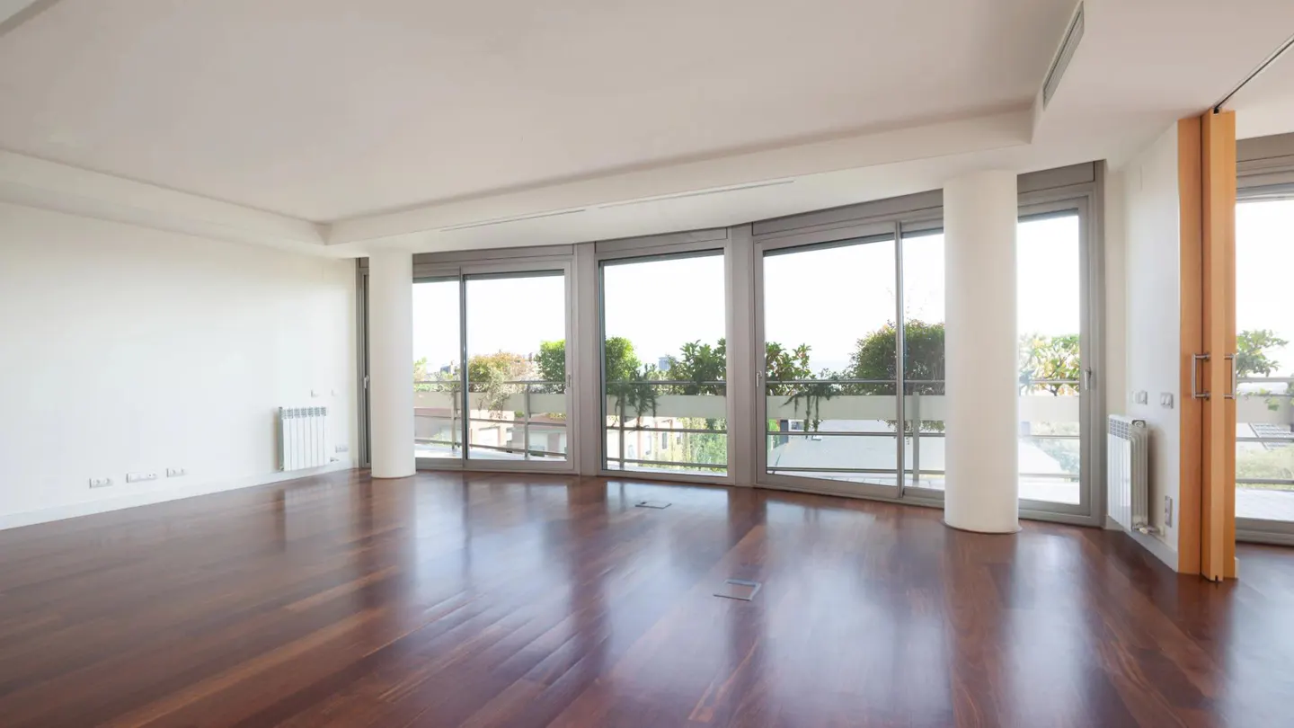 Bright, empty room with dark wood floors, white walls, and large windows overlooking greenery. Radiators are visible.