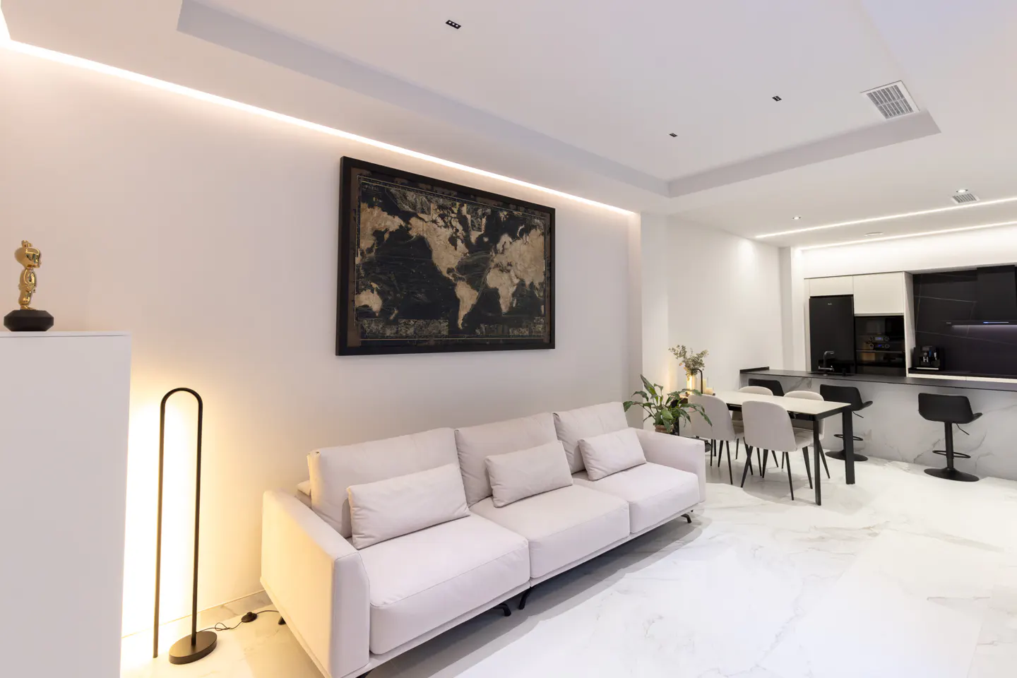 Bright, modern living room with a white sofa, marble floors, and a world map art piece. A dining table and kitchen bar are in the background.