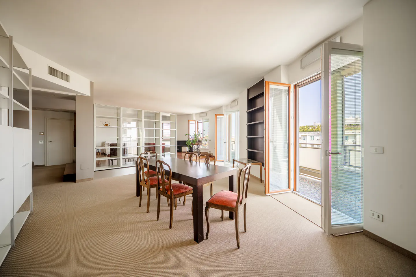 Bright, modern dining room with a dark wood table, red cushioned chairs, and open balcony doors. White shelving lines the walls.